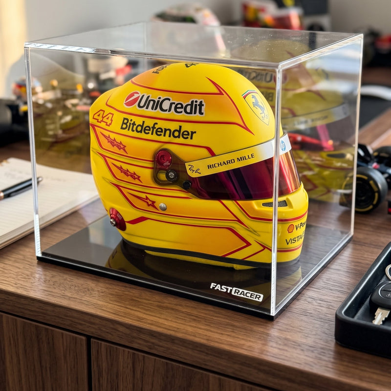 A yellow Formula 1 racing helmet with red and black graphics is displayed inside a clear acrylic case on a black base, set on a wooden desk.